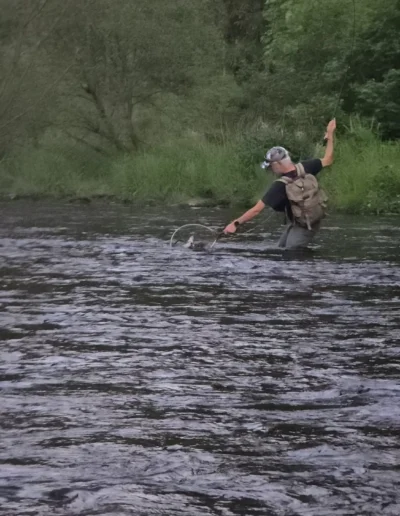 Man wading in a river with a fishing rod in hand, casting or snagging line while wearing a cap and backpack.