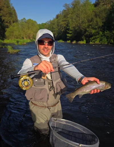 Man in a hooded jacket and cap standing in a river, proudly holding a large rainbow trout over a landing net.