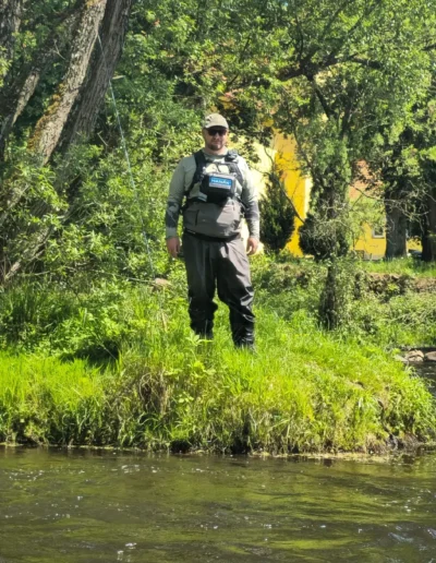 Man in waders and fishing vest stands on a grassy riverbank beside a flowing river, with trees and a yellow building in the background.