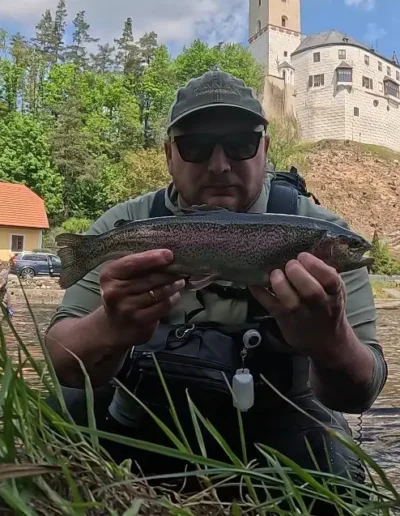 Man kneeling at a riverbank, holding a rainbow trout with a castle on a hill and red-roofed houses in the background.