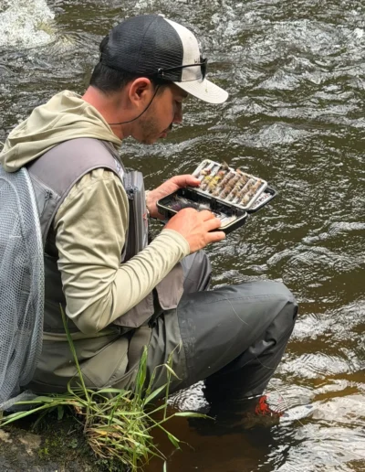 Fisherman in a cap and vest crouches at a riverbank, sorting a fly box with assorted flies for fishing.
