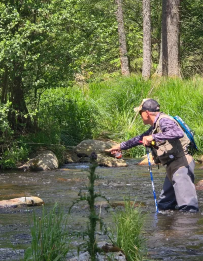 Man fishing in a shallow forest stream, wearing a cap and vest, casting his rod among rocks and grasses.