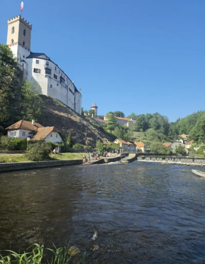 Castle Rosenberg in Czech Republic on a hill overlooking a river, with a white-walled fortress and tall tower topped by a flag, beside a tree-lined bank and village houses; a group of people stroll along the riverside path under a clear blue sky.