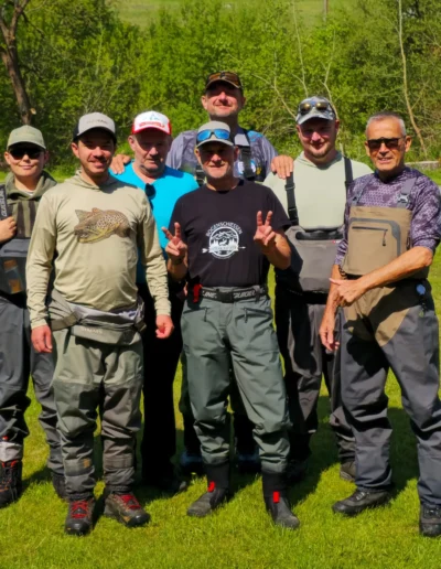 Seven flyfishers in outdoor gear standing on a grassy field with trees in the background, posing for a group photo.