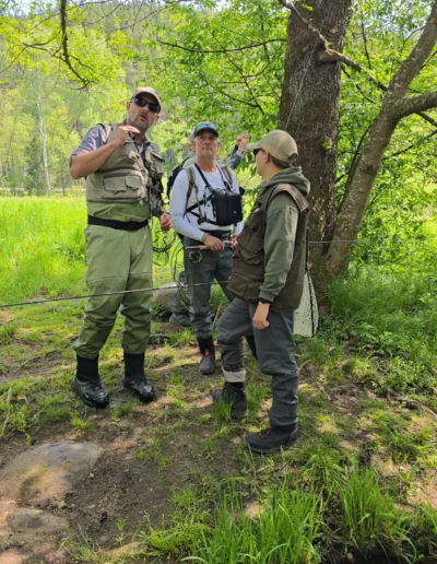 Workshop for flyfishers researchers in vests and backpacks stand by a tree along a grassy riverbank, discussing fieldwork gear.