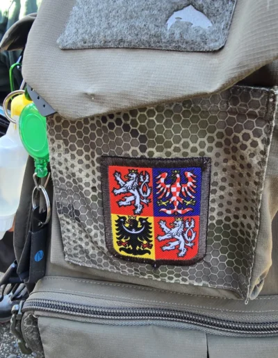 Flyfisher-style vest with hex-pattern fabric and a colorful 4-quadrant heraldic patch on the chest side.